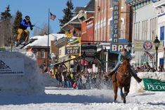 a person on a horse jumping over a snow bank in the middle of town with people watching