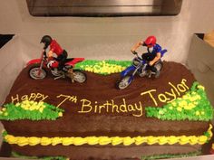 two motorcyclists on dirt bikes riding across a birthday cake in a box