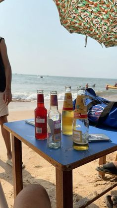 several bottles of soda sit on a table at the beach while people stand in the background