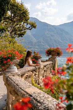 a woman is sitting on a balcony overlooking the water and mountains with red flowers in pots