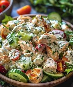 a wooden bowl filled with chicken salad next to tomatoes and cucumbers on a table