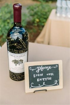 a bottle of wine sitting next to a chalkboard sign that reads please sign our bottle