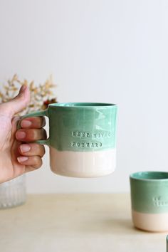 a person holding a green and white coffee mug next to another cup on a table