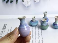 a hand holding a miniature vase in front of three other small vases on top of a book