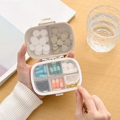 a person holding an open pill box on a table next to a glass of water