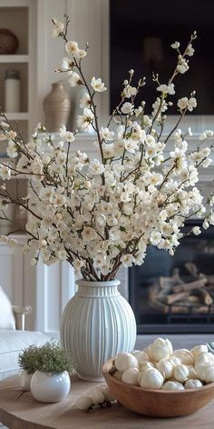 white flowers in a vase and some eggs on a table with a fireplace in the background