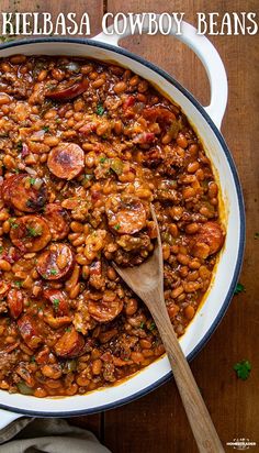 a large white pot filled with beans and sausages next to a wooden spoon on top of a table