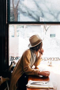 a woman sitting at a table in front of a window wearing a hat and looking out the window