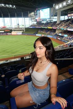 a beautiful young woman sitting on top of a blue chair in front of a baseball field