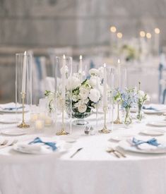 the table is set with white and blue flowers in vases, candles, and plates