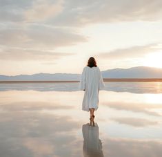 a woman walking across a large body of water with mountains in the background at sunset