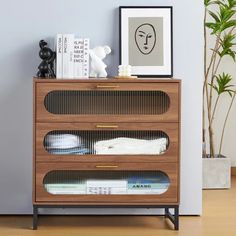 a wooden dresser with three drawers and pictures on the wall above it, next to a potted plant