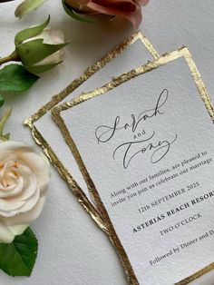 two wedding cards with gold foil on them next to a white rose and green leaves