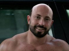 a bald man with no shirt standing in front of a car