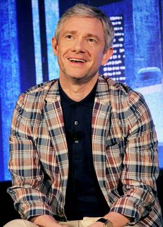 a man sitting in front of a blue screen with his hands crossed and looking at the camera