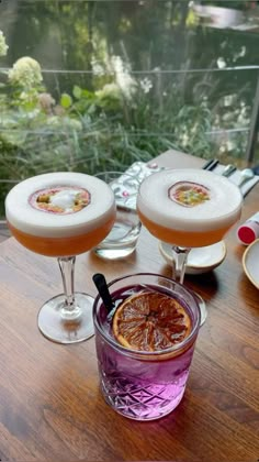 three glasses filled with drinks sitting on top of a wooden table next to plates and utensils