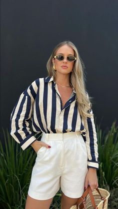 a woman in white shorts and striped shirt holding a straw bag with her hands on her hips