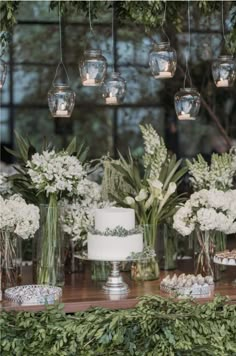 a table topped with lots of vases filled with white flowers and greenery next to a cake