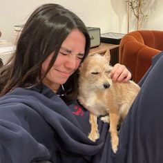 a woman holding a small dog wrapped in a blanket on her lap and smiling at the camera