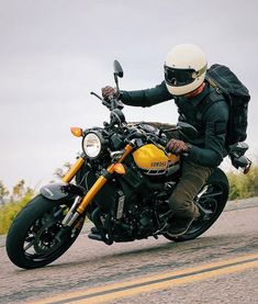 a man riding on the back of a yellow and black motorcycle down a street next to tall grass