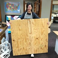 a woman holding up a large piece of plywood in an office area with other furniture