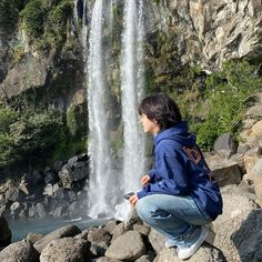 a woman sitting on rocks next to a waterfall and looking at something in the water