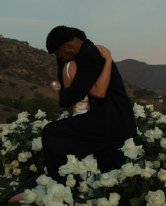 a man and woman are hugging in front of white flowers at sunset with mountains in the background