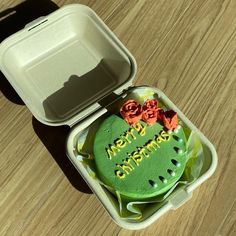 a green cake in a foam container on a wooden table next to a white box with the words merry christmas written on it