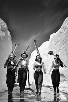 three women in bathing suits carrying skis and snowboards on their shoulders, walking through the snow