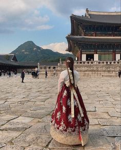 a woman wearing a red and white dress standing in front of a building with mountains behind her