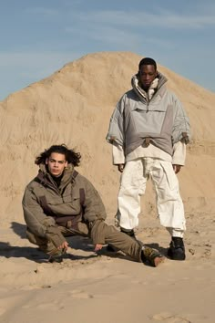 two men sitting on the sand in front of a large hill and one man standing next to him