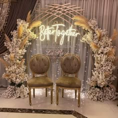 two chairs sitting in front of a wedding arch with lights and flowers on the wall