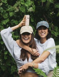 two women standing next to each other in front of some trees and bushes, one holding a coffee cup