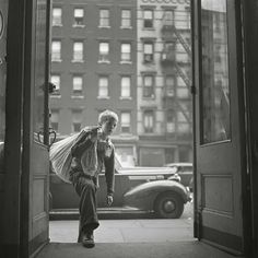 an old man walking out of a building with a bag on his back and cars parked in the background