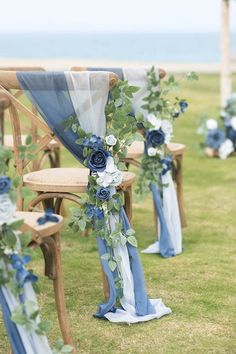 an outdoor ceremony with blue flowers and greenery on the aisle, along with wooden chairs