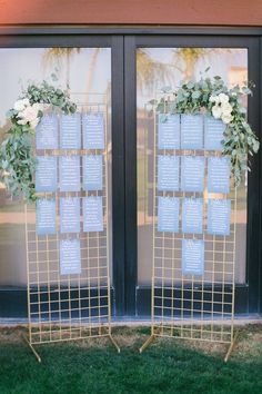 two gold metal stands with seating cards on them and flowers in front of the doors