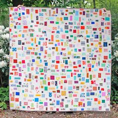 a colorful quilt is displayed in front of some trees and bushes with white flowers behind it