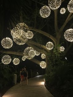 two people standing on a wooden walkway with lights hanging from the trees in the background