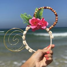 a hand holding up a flower and seashell headband on the beach with waves in the background