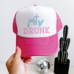 a pink and white trucker hat with the words just drunk on it next to cactus