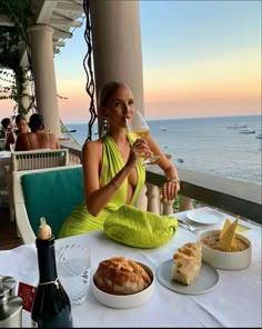 a woman sitting at a table with food and wine in front of the ocean while holding a glass of wine