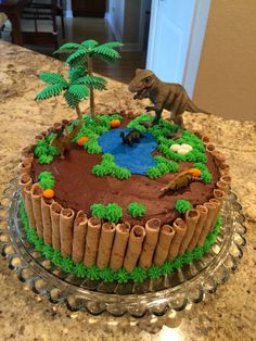 a dinosaur cake on a glass platter with trees and rocks in the middle,