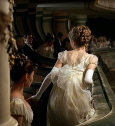 two women in white dresses and gloves are walking down the stairs at a formal event