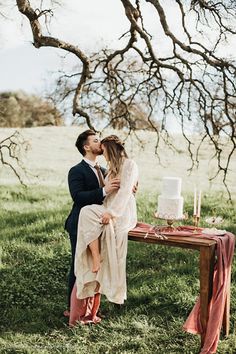 a man and woman kissing in front of a table with a cake on it, surrounded by trees