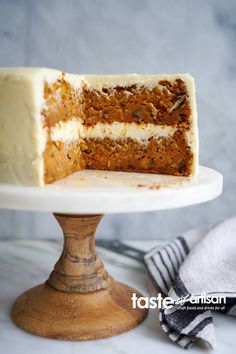 a carrot cake with white frosting on a wooden stand next to a slice missing from the cake