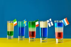 four glasses filled with different colored liquids on a yellow table next to a blue wall