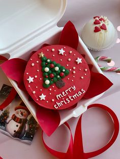 a decorated christmas cookie in a box next to a cupcake on a plate with a red ribbon