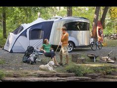 a man and woman standing next to a camper trailer with a dog laying on the ground