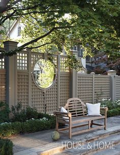 a wooden bench sitting under a tree next to a white fence and some bushes with flowers