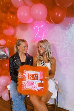 two women sitting next to each other in front of pink and orange balloons with the number 21 on it